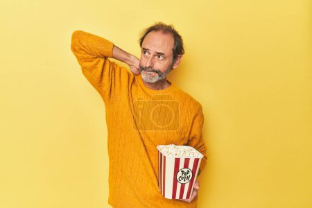 Photo for Man enjoying popcorn in yellow studio touching back of head, thinking and making a choice. - Royalty Free Image
