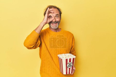 Téléchargez les photos : Homme appréciant pop-corn en studio jaune excité garder ok geste sur l'œil. - en image libre de droit