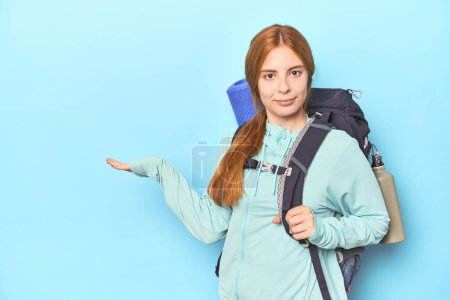 Photo for Redhead traveler with backpack and mat in studio showing a copy space on a palm and holding another hand on waist. - Royalty Free Image