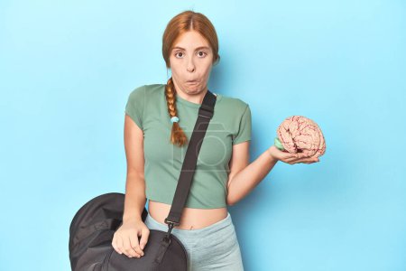 Photo for Sporty redhead with brain model on blue background shrugs shoulders and open eyes confused. - Royalty Free Image