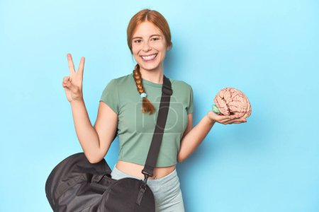 Photo for Sporty redhead with brain model on blue background joyful and carefree showing a peace symbol with fingers. - Royalty Free Image