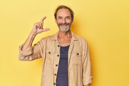 Photo for Caucasian middle-aged man in yellow studio holding something little with forefingers, smiling and confident. - Royalty Free Image