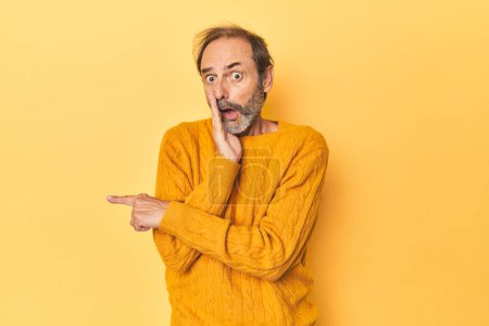 Photo for Caucasian middle-aged man in yellow studio saying a gossip, pointing to side reporting something. - Royalty Free Image