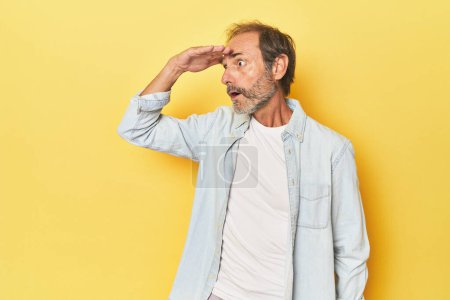 Photo for Caucasian middle-aged man in yellow studio looking far away keeping hand on forehead. - Royalty Free Image