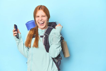 Photo for Mountaineer redhead with phone and backpack in blue studio - Royalty Free Image