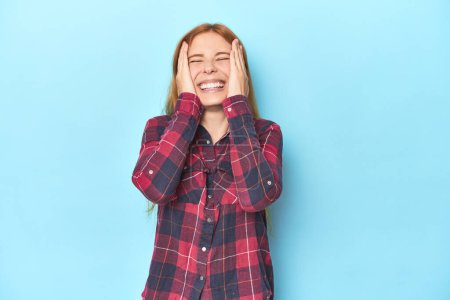 Photo for Redhead young woman on blue background laughs joyfully keeping hands on head. Happiness concept. - Royalty Free Image