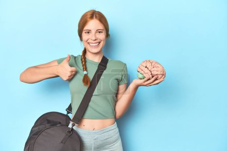 Photo for Sporty redhead with brain model on blue background smiling and raising thumb up - Royalty Free Image