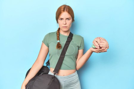 Photo for Sporty redhead with brain model on blue background screaming very angry and aggressive. - Royalty Free Image
