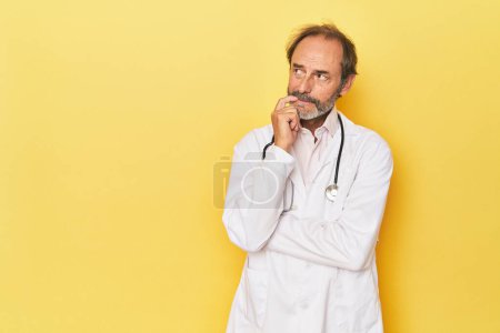 Foto de Doctor con estetoscopio en estudio amarillo relajado pensando en algo mirando un espacio de copia. - Imagen libre de derechos