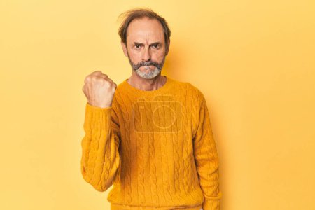 Téléchargez les photos : Blanc homme d'âge moyen en studio jaune montrant poing à caméra, expression faciale agressive. - en image libre de droit