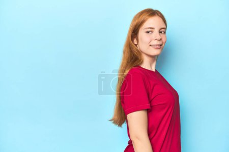 Photo for Redhead young woman on blue background looks aside smiling, cheerful and pleasant. - Royalty Free Image