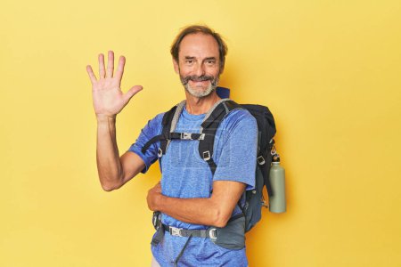 Photo for Middle-aged hiker with backpack in studio smiling cheerful showing number five with fingers. - Royalty Free Image