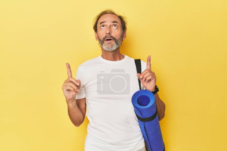 Téléchargez les photos : Homme avec tapis de yoga en studio jaune pointant à l'envers avec bouche ouverte. - en image libre de droit