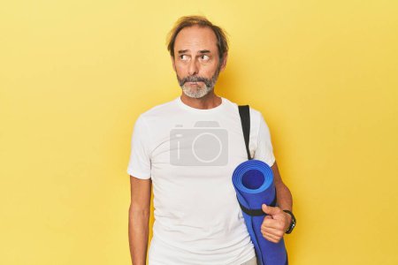 Téléchargez les photos : Homme avec tapis de yoga en studio jaune confus, se sent douteux et incertain. - en image libre de droit