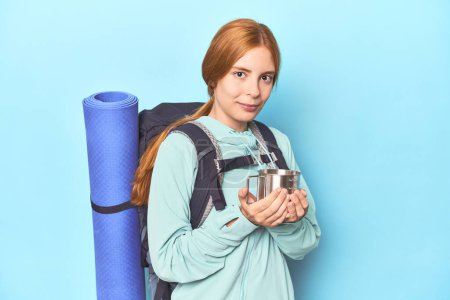 Photo for Mountaineer redhead sipping from camping cup in blue studio - Royalty Free Image