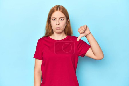 Téléchargez les photos : Jeune femme rousse sur fond bleu montrant un geste détestable, pouces baissés. Concept de désaccord. - en image libre de droit