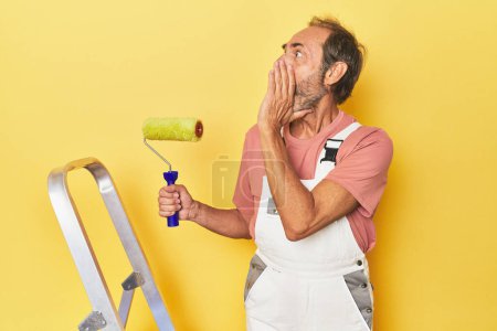 Photo for Man painting yellow backdrop in studio shouting and holding palm near opened mouth. - Royalty Free Image