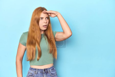Photo for Redhead young woman on blue background looking far away keeping hand on forehead. - Royalty Free Image