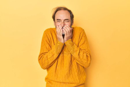 Foto de Caucásico hombre de mediana edad en estudio amarillo riendo de algo, cubriendo la boca con las manos. - Imagen libre de derechos