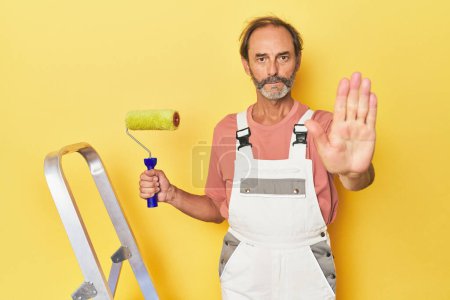 Photo for Man painting yellow backdrop in studio standing with outstretched hand showing stop sign, preventing you. - Royalty Free Image