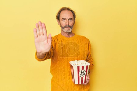 Téléchargez les photos : Homme profitant de pop-corn en studio jaune debout avec la main tendue montrant le panneau d'arrêt, vous empêchant. - en image libre de droit