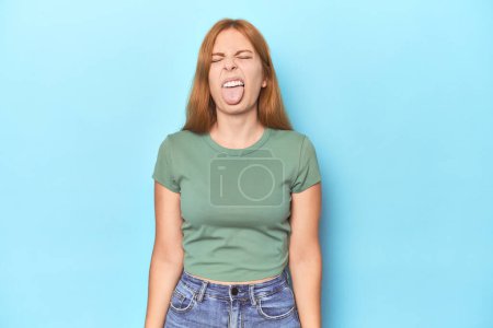 Téléchargez les photos : Rousse jeune femme sur fond bleu drôle et amical collant sur la langue. - en image libre de droit