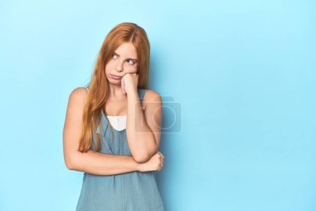 Téléchargez les photos : Rousse jeune femme sur fond bleu qui se sent triste et coûteux, en regardant l'espace de copie. - en image libre de droit