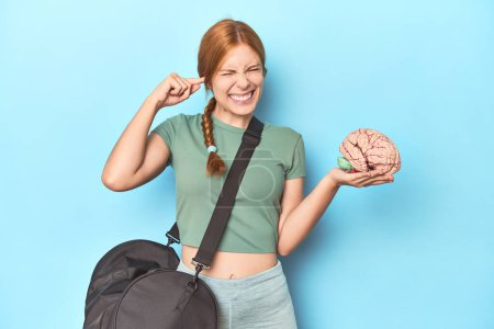 Photo for Sporty redhead with brain model on blue background covering ears with hands. - Royalty Free Image