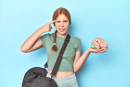 Photo for Sporty redhead with brain model on blue background showing a disappointment gesture with forefinger. - Royalty Free Image