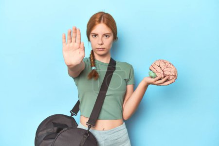 Photo for Sporty redhead with brain model on blue background standing with outstretched hand showing stop sign, preventing you. - Royalty Free Image