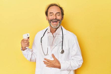 Foto de Doctor sosteniendo termómetro infrarrojo en el estudio se ríe en voz alta manteniendo la mano en el pecho. - Imagen libre de derechos