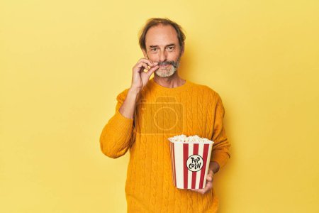 Photo for Man enjoying popcorn in yellow studio with fingers on lips keeping a secret. - Royalty Free Image