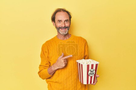 Téléchargez les photos : Homme appréciant le pop-corn en studio jaune souriant et pointant du doigt, montrant quelque chose à vide. - en image libre de droit