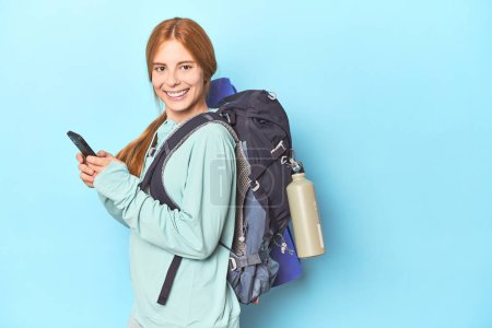 Photo for Mountaineer redhead with phone and backpack in blue studio - Royalty Free Image