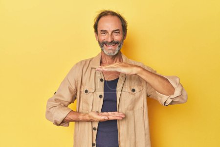 Foto de Caucásico hombre de mediana edad en estudio amarillo sosteniendo algo con ambas manos, presentación del producto. - Imagen libre de derechos