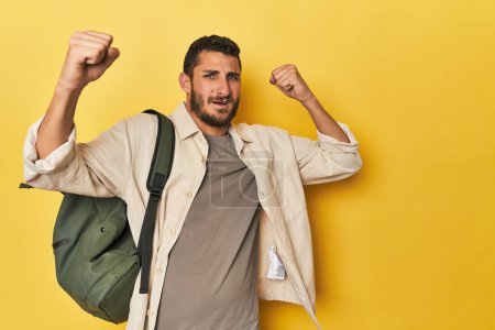Photo for Young Hispanic man with travel backpack raising fist after a victory, winner concept. - Royalty Free Image