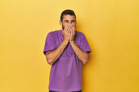 Foto de Joven hispano sobre fondo amarillo riéndose de algo, cubriendo la boca con las manos. - Imagen libre de derechos