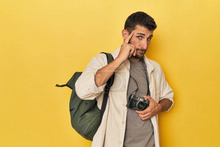Photo for Young Hispanic travel photographer poses pointing temple with finger, thinking, focused on a task. - Royalty Free Image