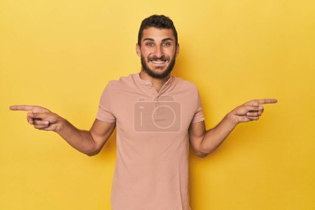 Téléchargez les photos : Jeune homme hispanique sur fond jaune pointant vers différents espaces de copie, en choisissant l'un d'eux, montrant avec le doigt. - en image libre de droit