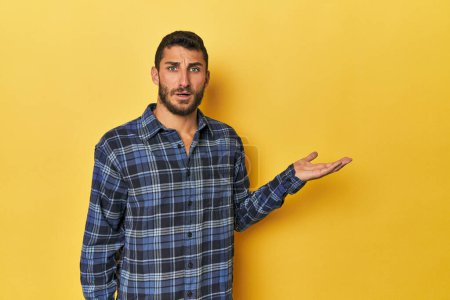 Photo for Young Hispanic man on yellow background impressed holding copy space on palm. - Royalty Free Image
