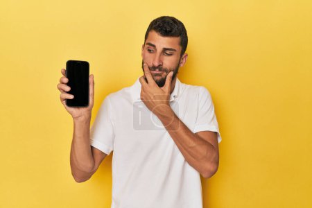 Photo for Young Hispanic man shows phone screen looking sideways with doubtful and skeptical expression. - Royalty Free Image