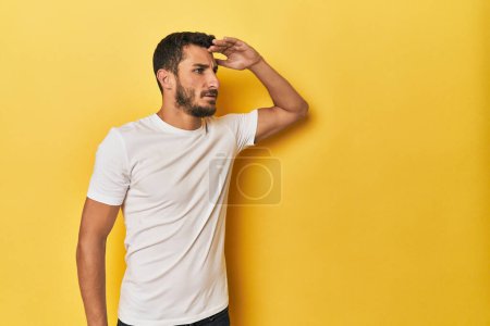 Foto de Joven hispano sobre fondo amarillo mirando lejos manteniendo la mano en la frente. - Imagen libre de derechos