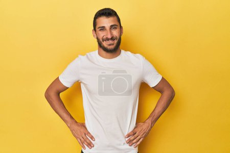 Téléchargez les photos : Jeune homme hispanique sur fond jaune confiant garder les mains sur les hanches. - en image libre de droit