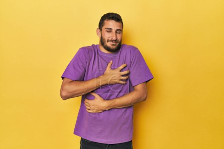 Foto de Joven hispano sobre fondo amarillo se ríe felizmente y se divierte manteniendo las manos en el estómago. - Imagen libre de derechos