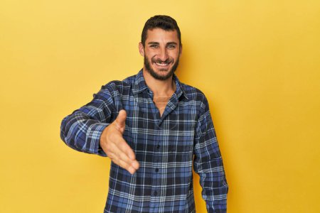 Téléchargez les photos : Jeune homme hispanique sur fond jaune tendre la main à la caméra dans le geste de salutation. - en image libre de droit