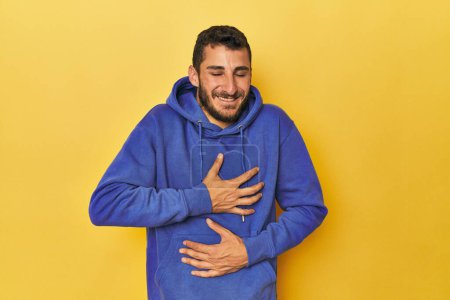 Photo for Young Hispanic man on yellow background laughs happily and has fun keeping hands on stomach. - Royalty Free Image