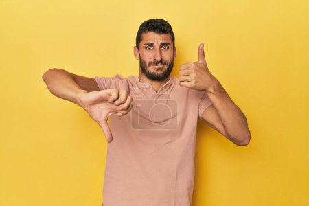 Téléchargez les photos : Jeune homme hispanique sur fond jaune montrant pouces vers le haut et pouces vers le bas, concept de choix difficile - en image libre de droit