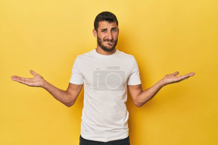 Téléchargez les photos : Jeune homme hispanique sur fond jaune épaules confuses et douteuses haussant pour tenir un espace de copie. - en image libre de droit
