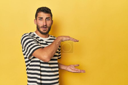 Foto de Joven hispano sobre fondo amarillo conmocionado y sorprendido sosteniendo un espacio de copia entre manos. - Imagen libre de derechos