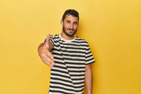 Téléchargez les photos : Jeune homme hispanique sur fond jaune tendre la main à la caméra dans le geste de salutation. - en image libre de droit